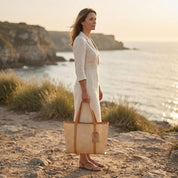 A woman in a white dress stands on a rocky cliff overlooking the ocean, holding a large woven bag.