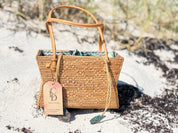 A woven straw handbag with a tag attached, placed on a sandy beach.