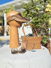 a wooden headband, a woven straw handbag, sunglasses, and a wooden stand with a tag attached, all placed on a white table.