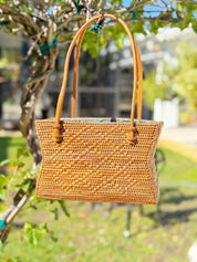 A woven straw handbag is hanging from a tree branch in a garden setting.