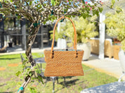 A woven brown purse is hanging from a tree branch in a lush, green outdoor setting.