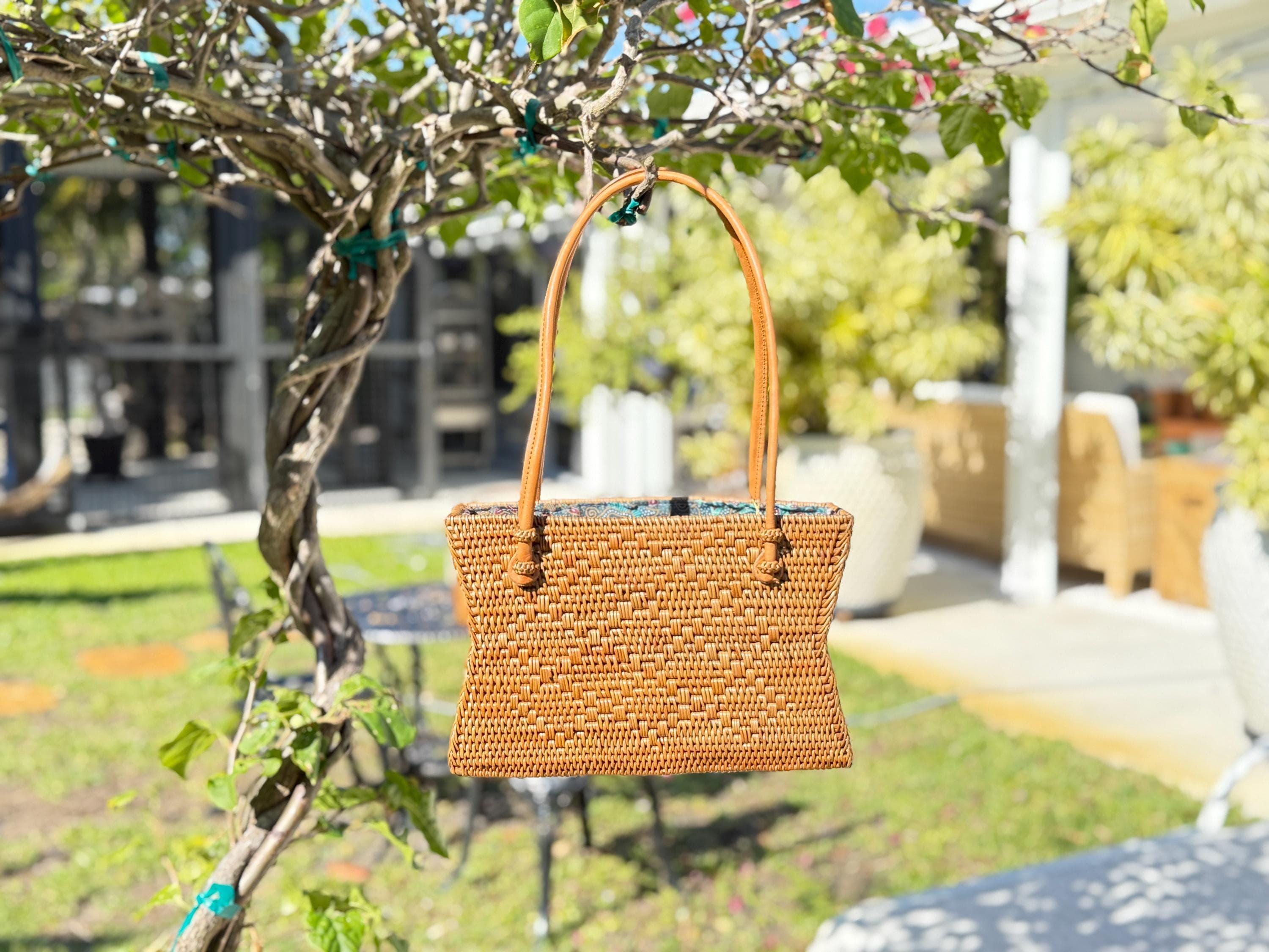 A woven brown purse is hanging from a tree branch in a lush, green outdoor setting.