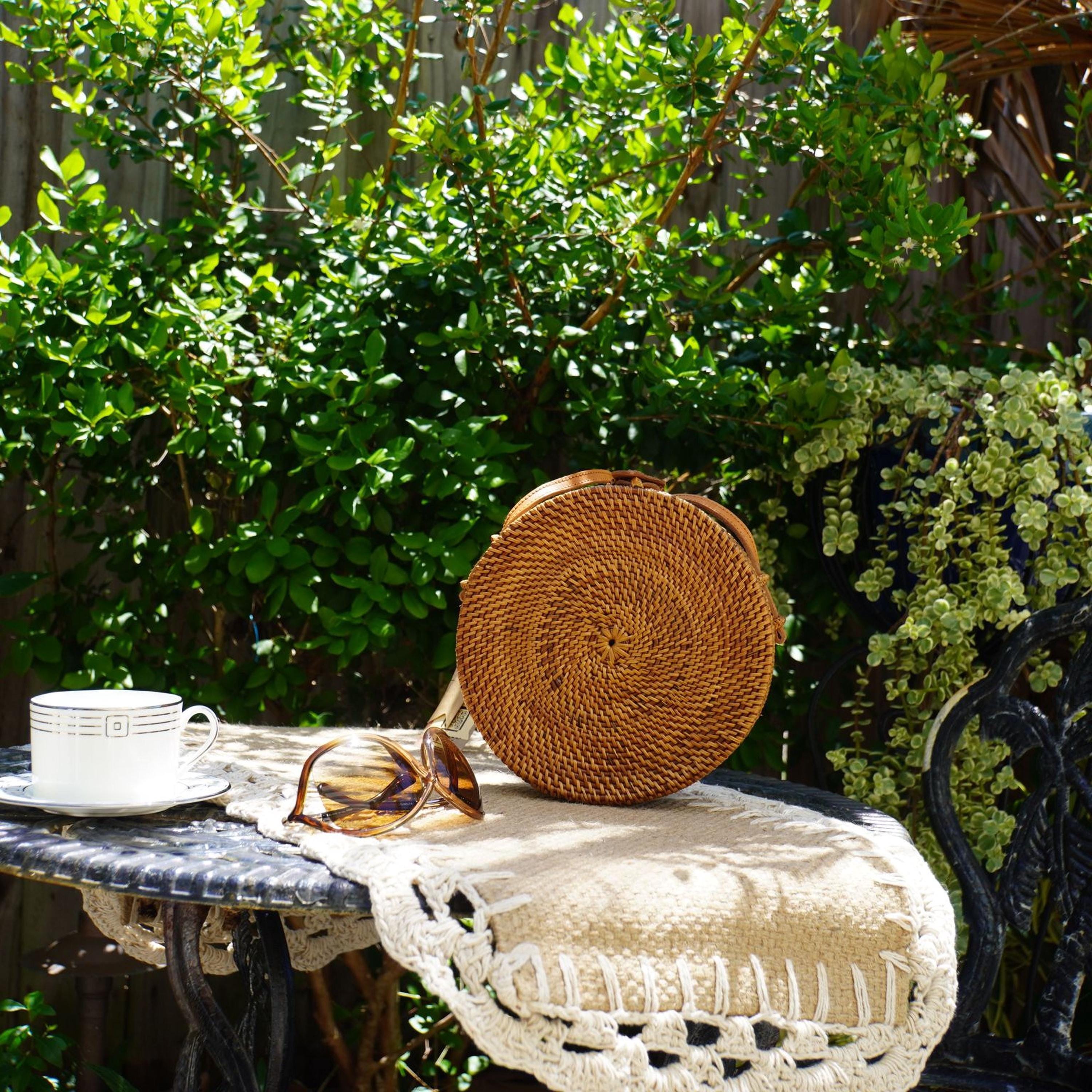 A round woven bag sits on a lace doily on a small table, accompanied by a white coffee cup and a pair of sunglasses, with a lush green garden and a black wrought iron chair in the background.