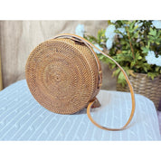 A round, brown woven bag with a strap is placed on a white surface, with a potted plant in the background.