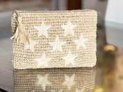 A beige woven bag with white star-shaped embellishments, sitting on a reflective surface.