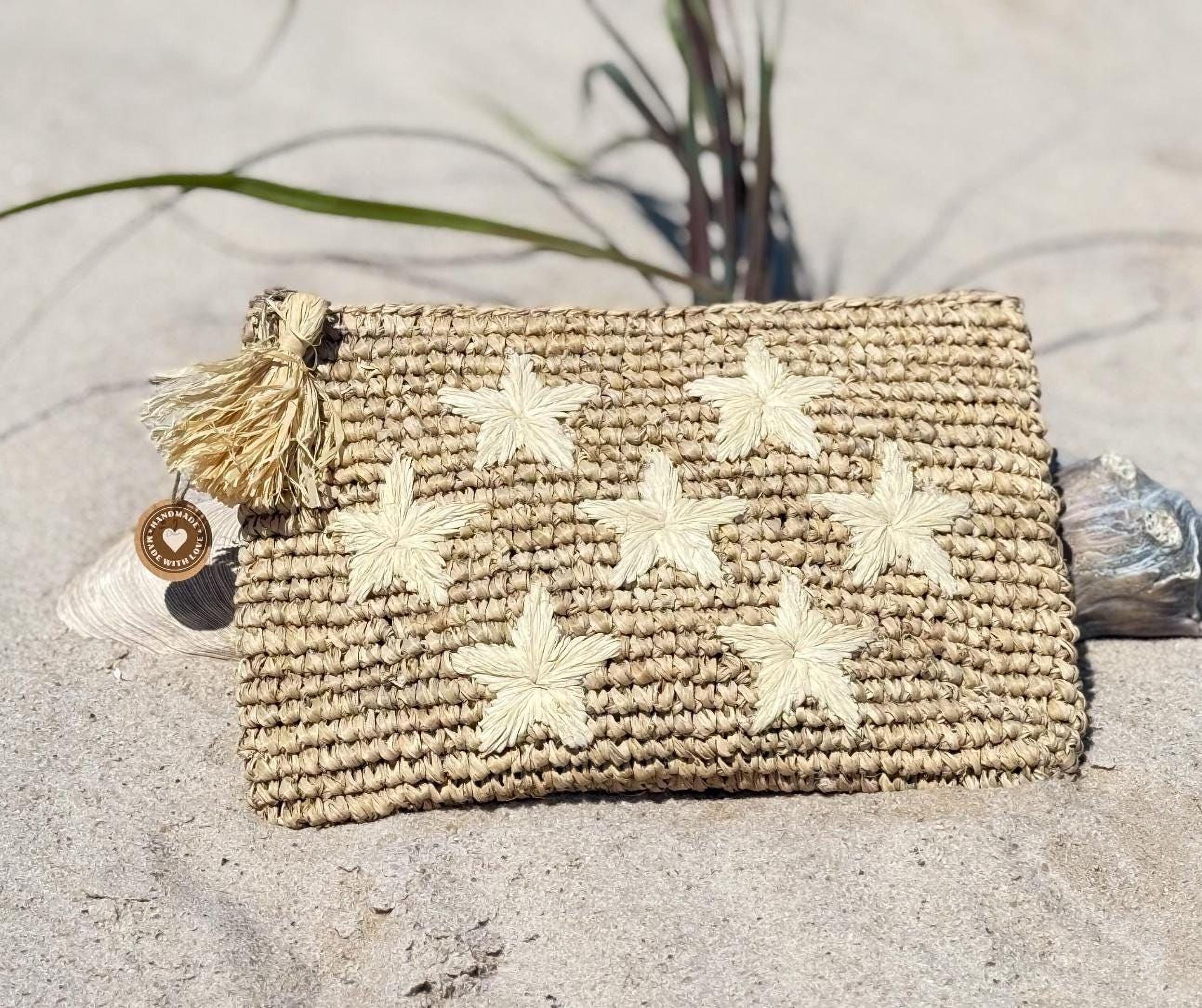 A woven clutch purse with white star-shaped embellishments rests on a sandy beach, with a small tassel attached to the side.
