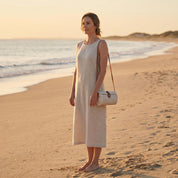 A woman in a white dress stands on a sandy beach, gazing out at the ocean at sunset.