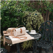A woven purse and a white coffee cup sit on a small table in a lush garden setting, surrounded by greenery and a black wrought iron chair.