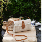 A beige woven handbag with a leather strap sits on a woven placemat, accompanied by a straw hat and a book, all placed on a metal table.