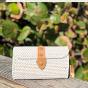 A white woven purse with a leather strap is placed on a wooden surface, with a blurred background of green leaves.