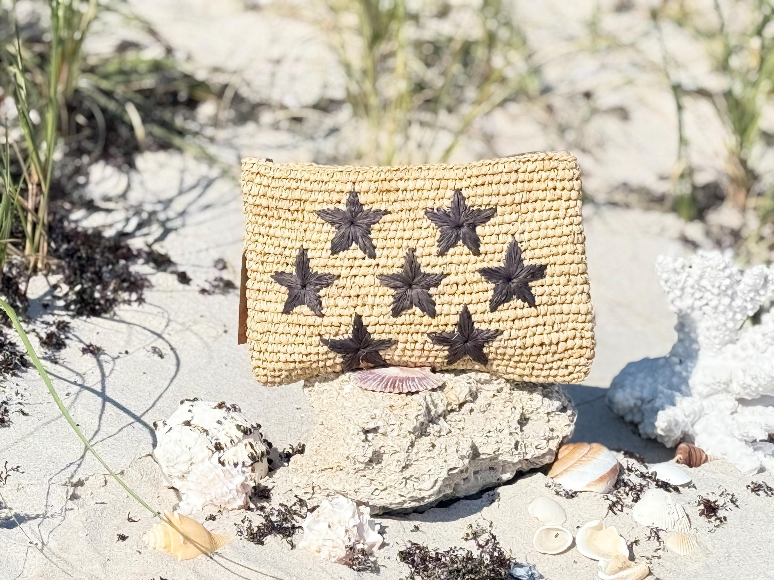 A woven straw bag with star-shaped embellishments sits on a rock in a sandy beach setting, surrounded by seashells and other natural elements.