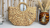 a woven straw bag with a round handle, placed on a white surface next to a vase filled with dried flowers.