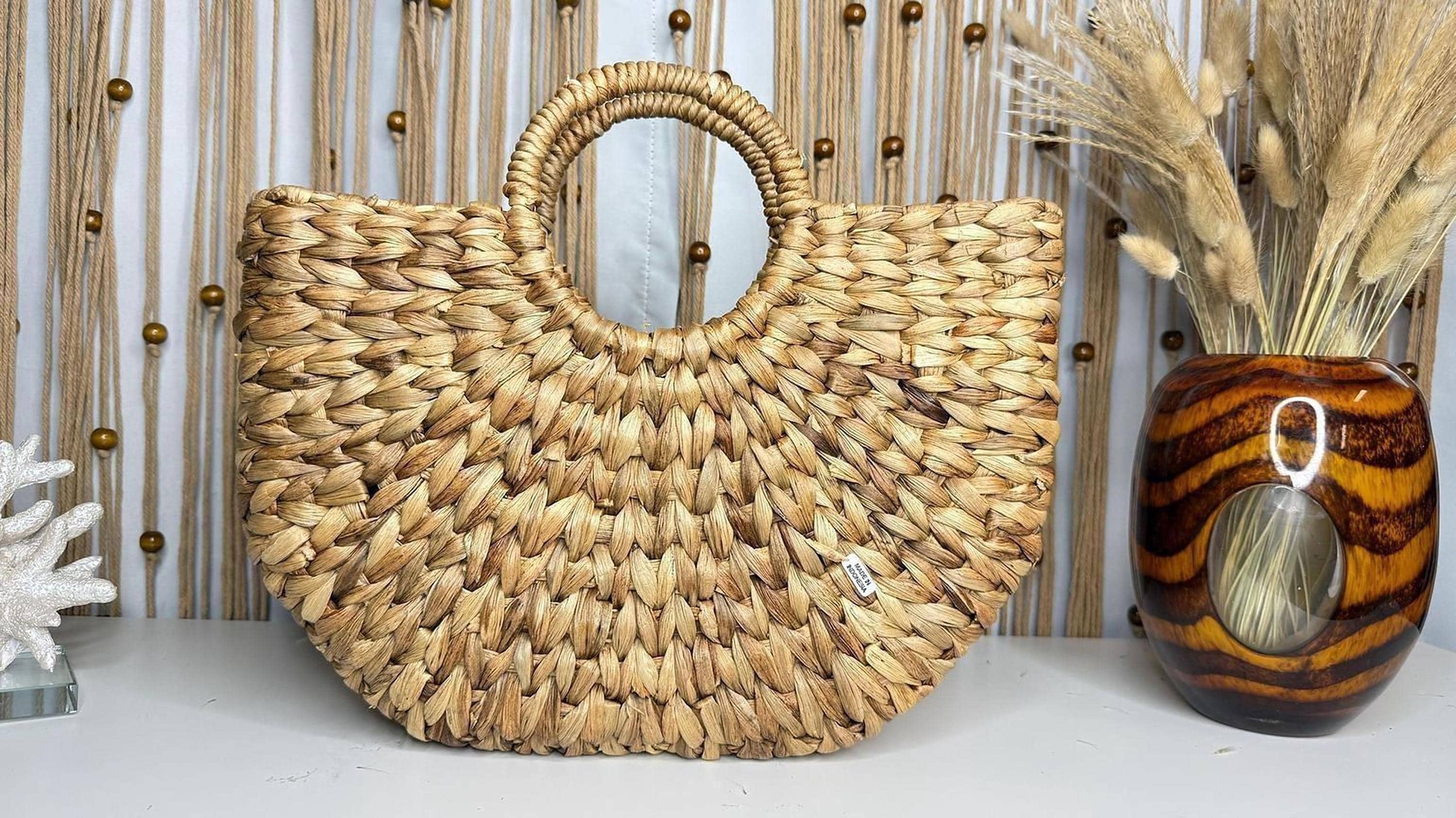 a woven straw bag with a round handle, placed on a white surface next to a vase filled with dried flowers.