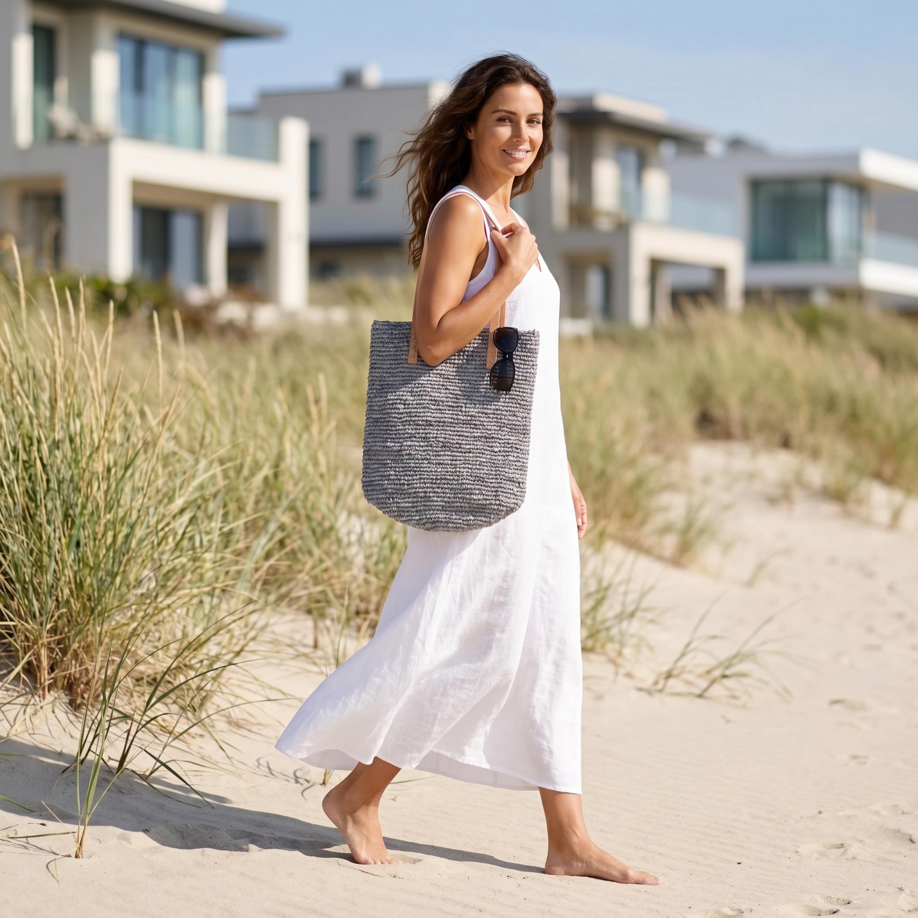A woman in a white dress walks along a sandy beach, carrying a large woven bag.