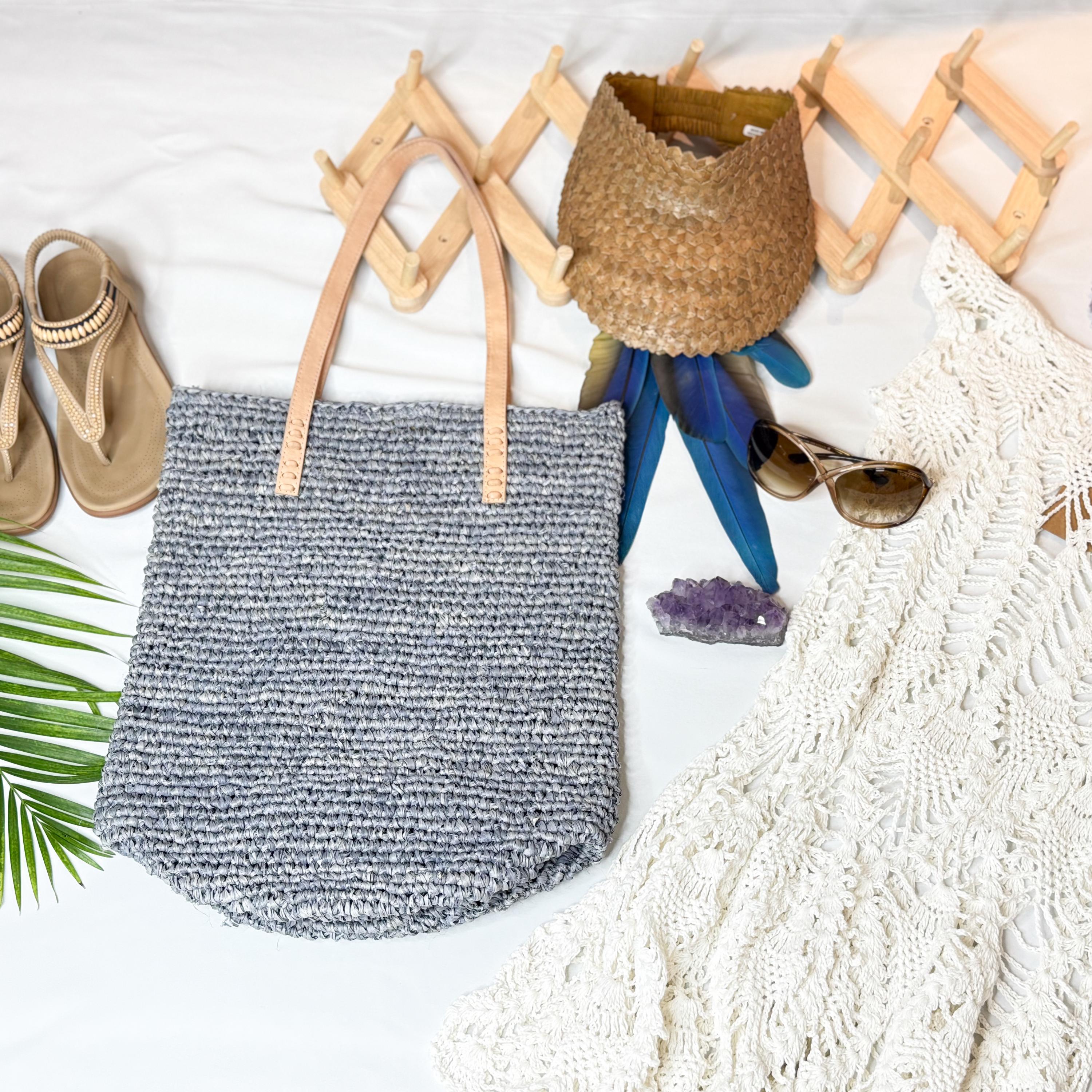 A handbag, a pair of sunglasses, a white lace garment, and a woven hat are arranged on a white surface, along with some natural elements like a green leaf.