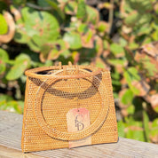 A woven straw handbag with a round handle and a tag displaying the letters "CD" is placed on a wooden surface against a blurred green background.