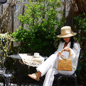 A woman in white sitting on a chair, holding a woven purse, with a small table and a cup of coffee in front of her.