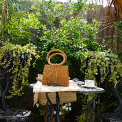 A woven handbag sits on a small table in a lush garden setting, surrounded by potted plants and a wooden fence.