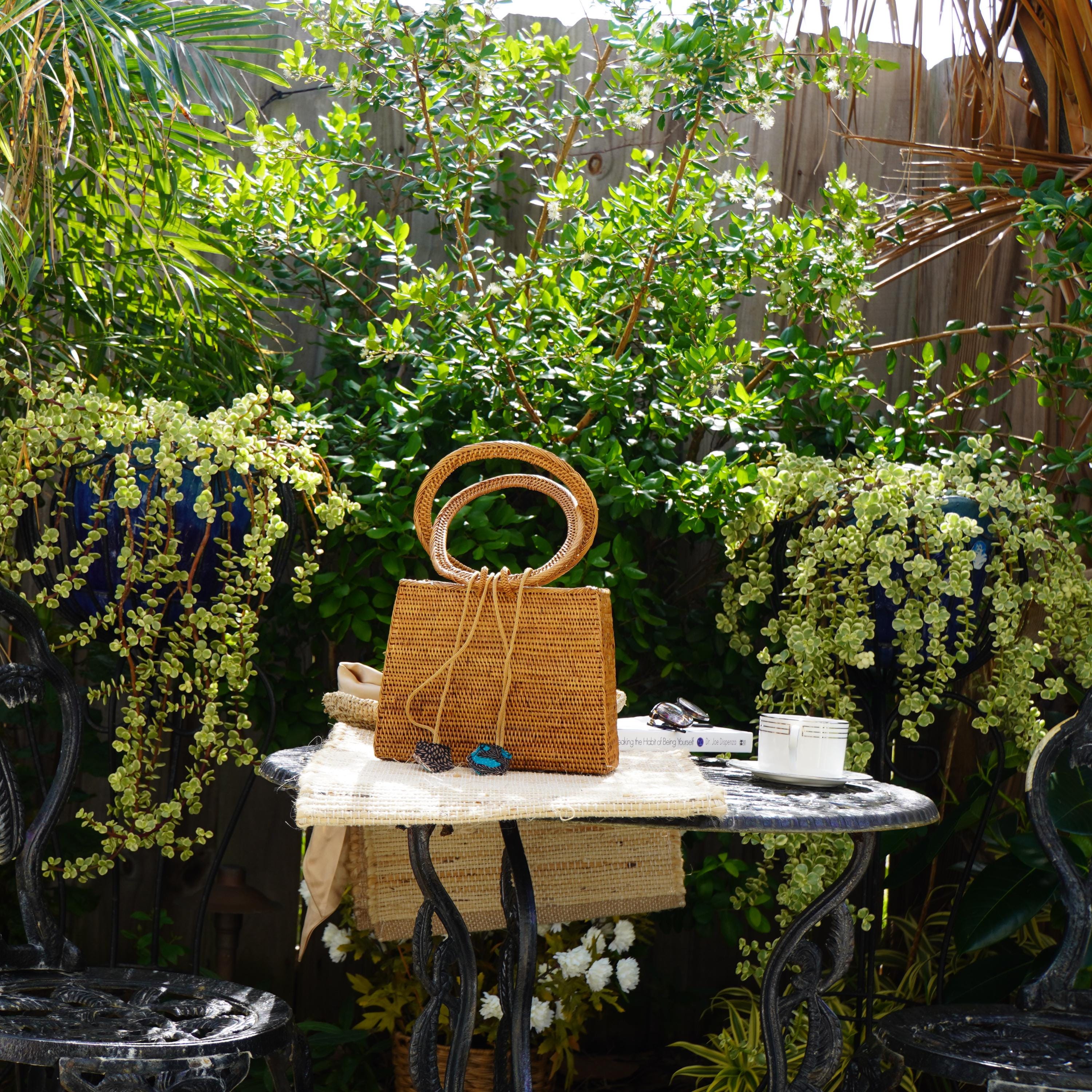 A woven handbag sits on a small table in a lush garden setting, surrounded by potted plants and a wooden fence.