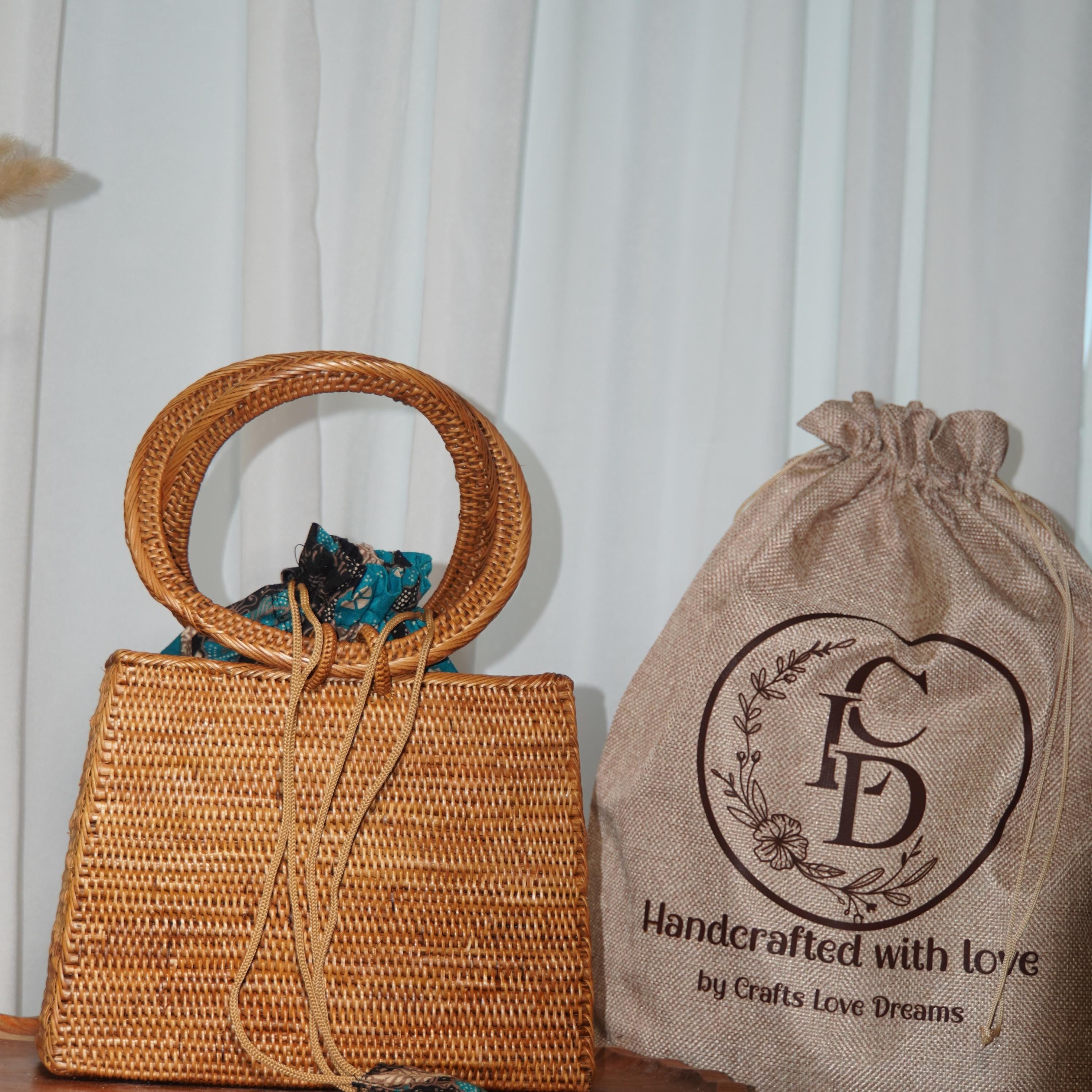 a woven handbag and a burlap bag with a logo, both placed on a white surface against a white curtain background.