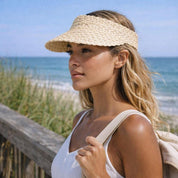 A woman wearing a straw hat and carrying a backpack stands near a wooden railing overlooking the ocean.