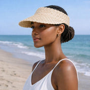 A woman wearing a straw hat and a white tank top stands on a beach, gazing out at the ocean.