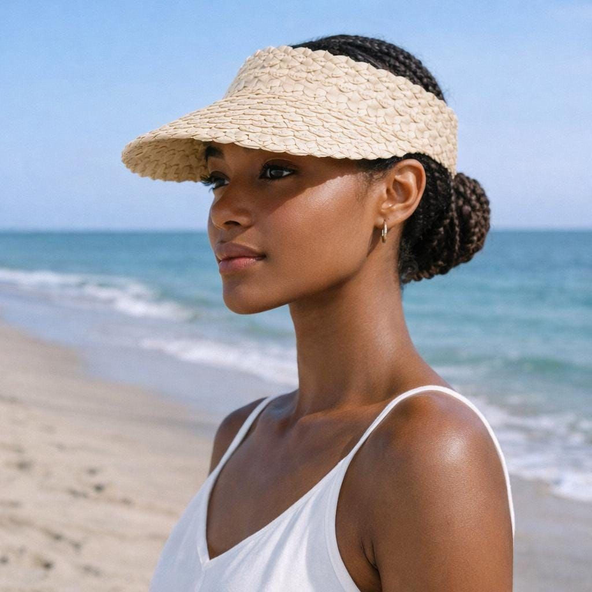 A woman wearing a straw hat and a white tank top stands on a beach, gazing out at the ocean.