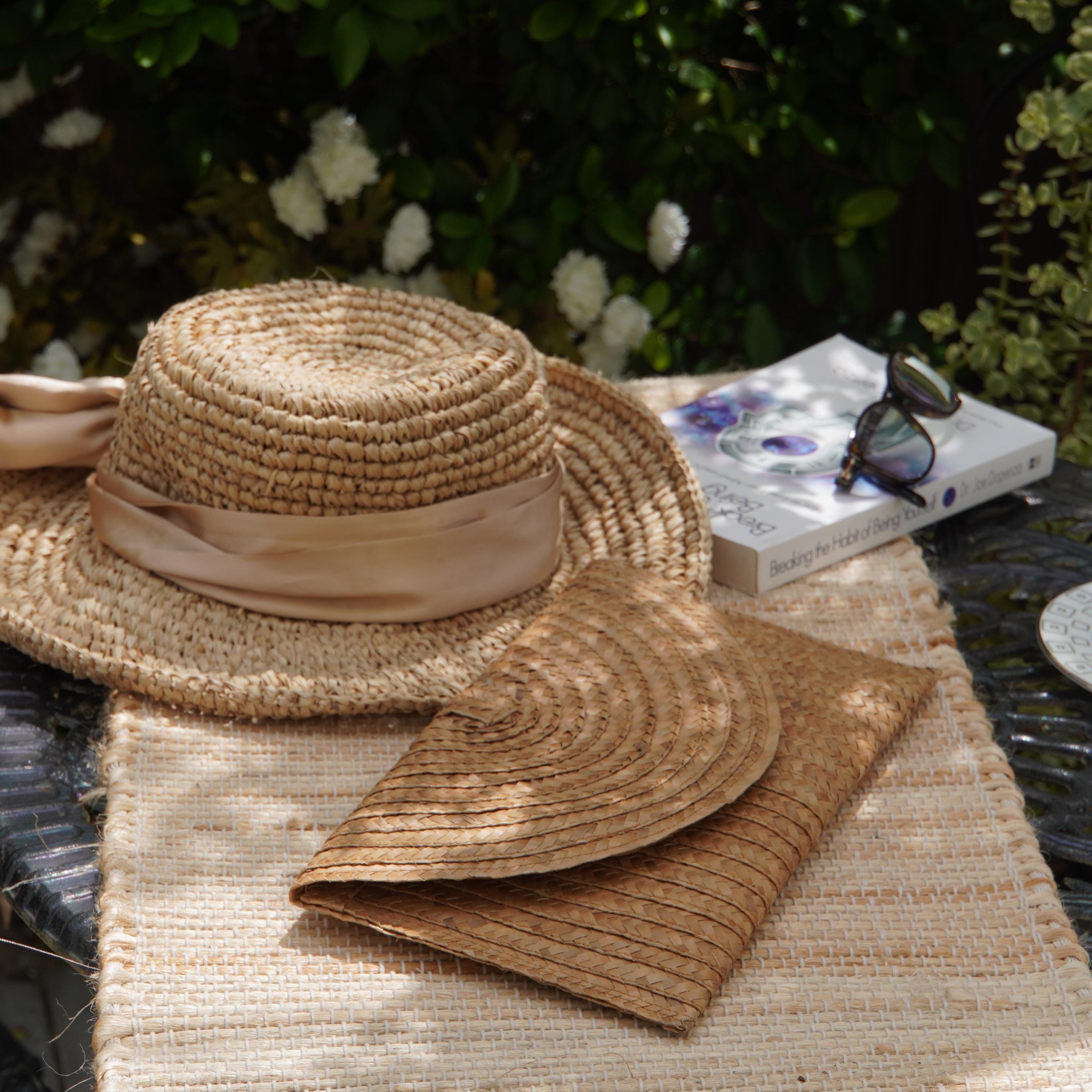 A straw hat, a book, and a small purse are arranged on a woven placemat, with a lush garden and white flowers in the background.