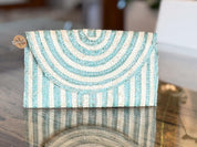 A light blue and white striped clutch purse is displayed on a reflective surface, with a tag attached to it.