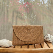 A brown woven purse is placed on a wooden surface, with a pink flower and a seashell visible in the background.