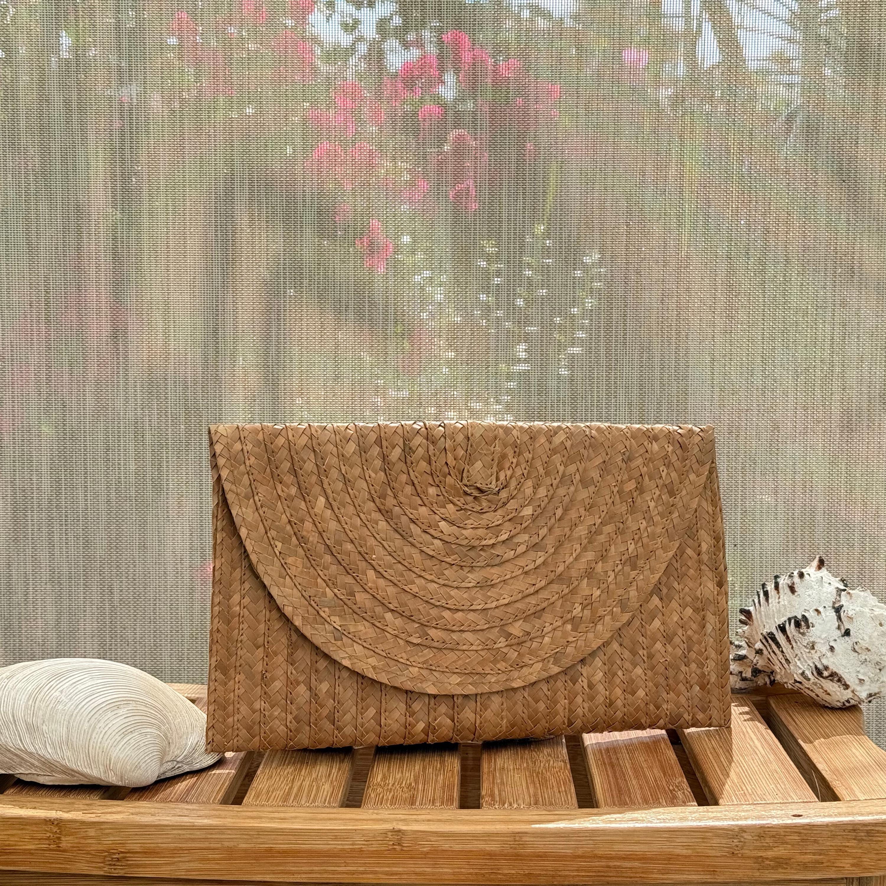 A brown woven purse is placed on a wooden surface, with a pink flower and a seashell visible in the background.