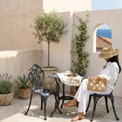 A woman in a white outfit is seated at a table with a woven bag, enjoying a drink in a potted plant and a small tree.