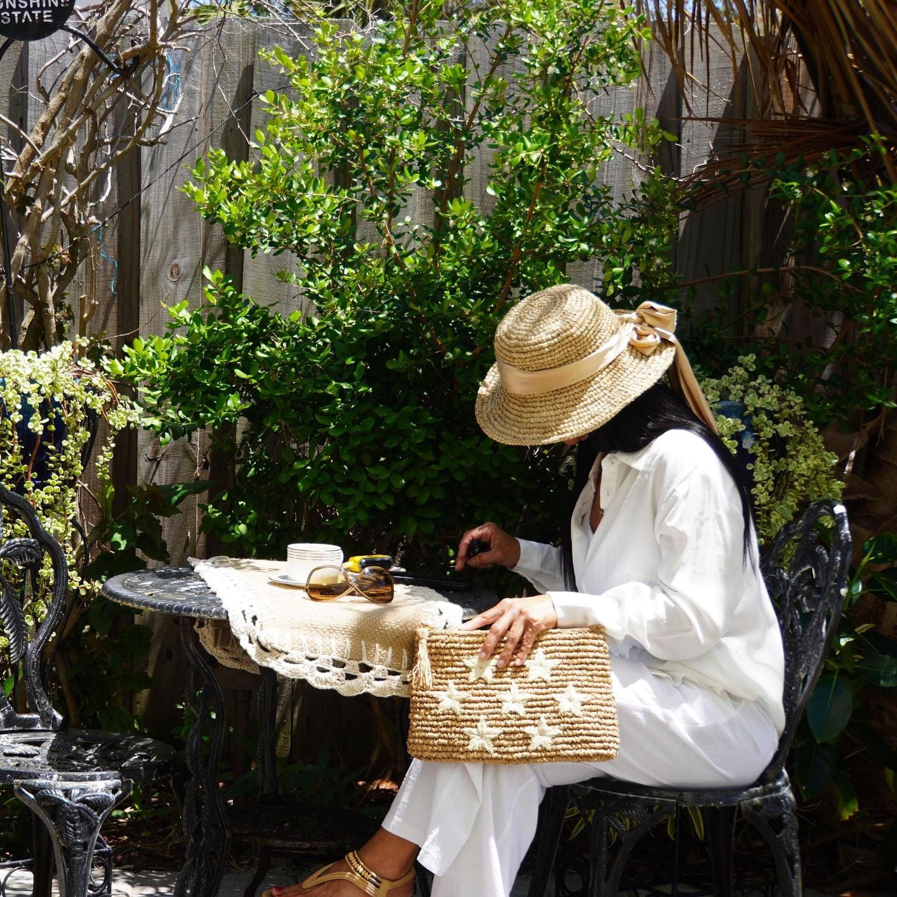 A woman in a white outfit and straw hat sits at a table with a woven bag, surrounded by lush greenery and a wooden fence.