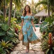 A woman in a blue and orange floral dress is walking on a stone path through a lush tropical garden, carrying a woven bag.