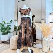 A mannequin wearing a black and orange tie-dye dress and matching skirt is displayed in a store, with a potted plant and a vase of flowers in the background.