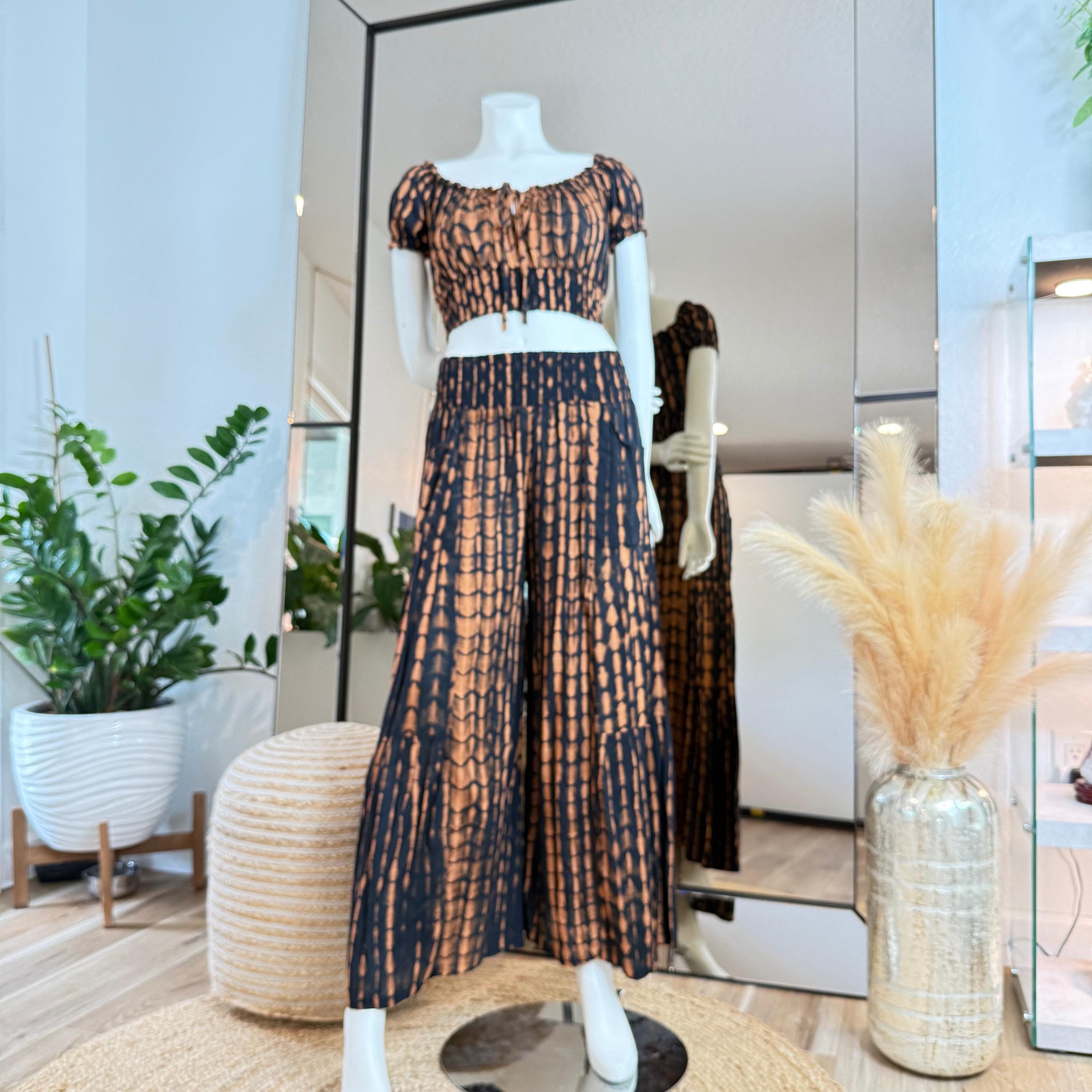 A mannequin wearing a black and orange tie-dye dress and matching skirt is displayed in a store, with a potted plant and a vase of flowers in the background.