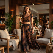 A woman in a black and orange patterned dress stands in a room with a mirror, holding a drink and surrounded by furniture.
