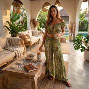 A woman in a green and white patterned outfit stands in a cozy, tropical-style room with a wooden table, potted plants, and a pool visible in the background.