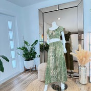 A mannequin wearing a green and white patterned dress stands in a room with a large mirror, surrounded by potted plants and a woven basket.