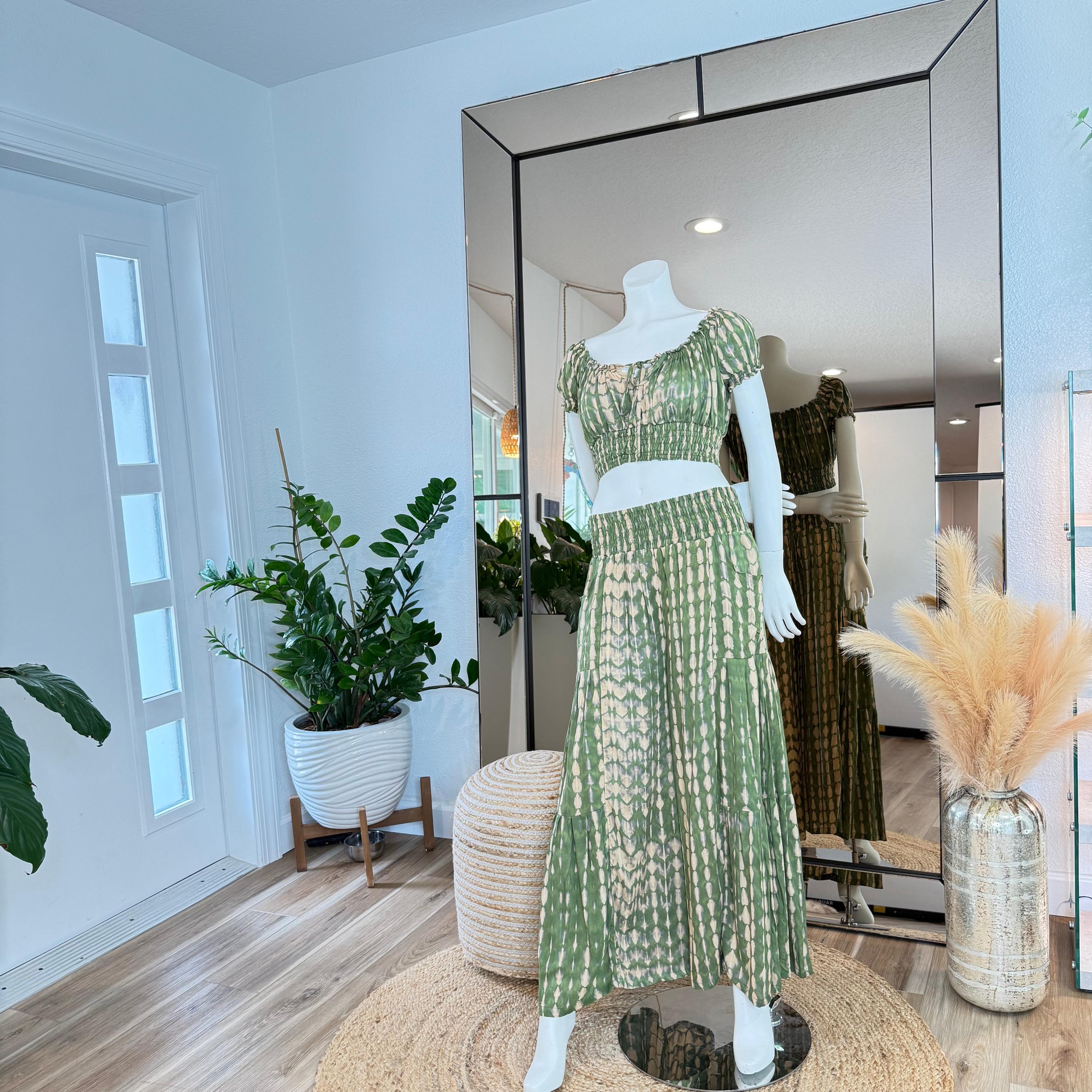 A mannequin wearing a green and white patterned dress stands in a room with a large mirror, surrounded by potted plants and a woven basket.