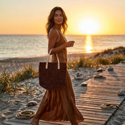 A woman in a brown dress walks along a wooden boardwalk on a beach at sunset, carrying a woven bag.