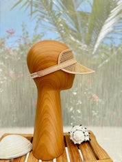 A wooden headless mannequin head wearing a beige woven visor stands on a wooden surface, with a small white object and a blurred background of greenery visible.