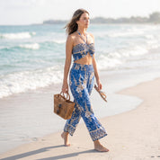 A woman in a blue and white patterned outfit walks along a sandy beach, carrying a brown woven bag.