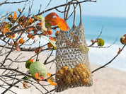 A woven mesh bag filled with yellow fruit hangs from a tree branch, with the bag suspended in the air against a backdrop of a sandy beach and the ocean.