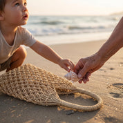 A young child reaches out to touch a woven straw bag on a sandy beach, with an adult's hand holding a seashell nearby.