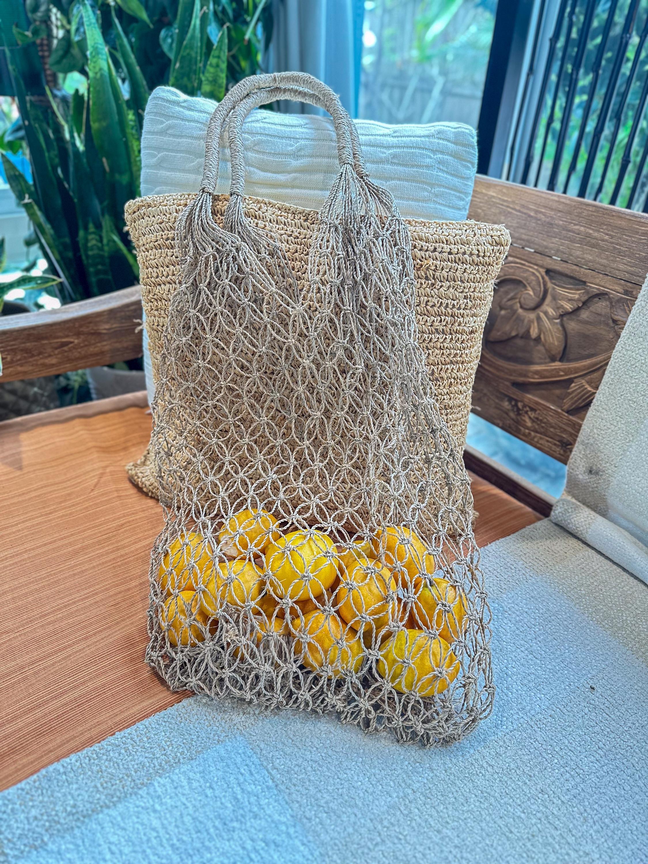 A woven mesh bag filled with bright yellow lemons sits on a wooden surface, with a wooden chair and a white pillow visible in the background.
