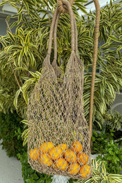 A woven mesh bag filled with bright yellow lemons is hanging from a rope, surrounded by lush green foliage.