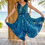 A woman in a blue and white tie-dye dress is posing on a wooden deck, with palm trees and the ocean visible in the background.