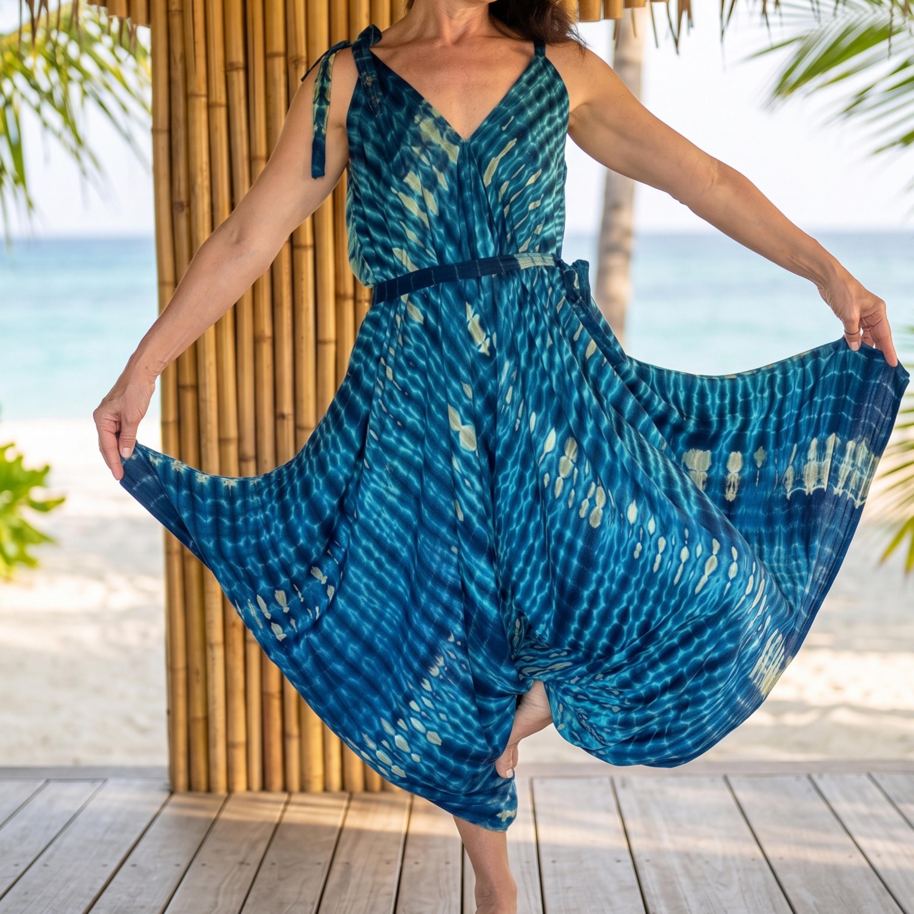 A woman in a blue and white tie-dye dress is posing on a wooden deck, with palm trees and the ocean visible in the background.