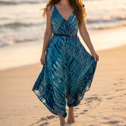A woman in a blue and white tie-dye dress walks along a sandy beach during sunset.
